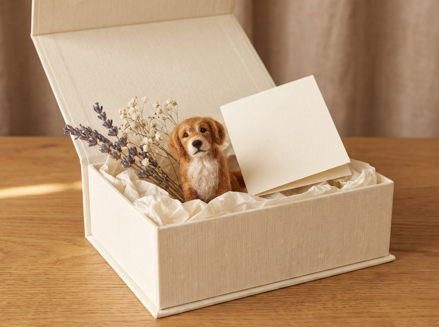 A cream-colored fabric gift box sits open on a wooden table, filled with tissue paper, dried lavender, and a small plush puppy toy. A blank cream card rests inside the box against a soft beige curtain background.