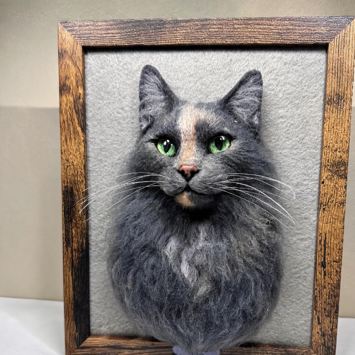 A framed portrait of a fluffy gray cat with bright green eyes, mounted on a textured gray background inside a rustic dark wood frame.